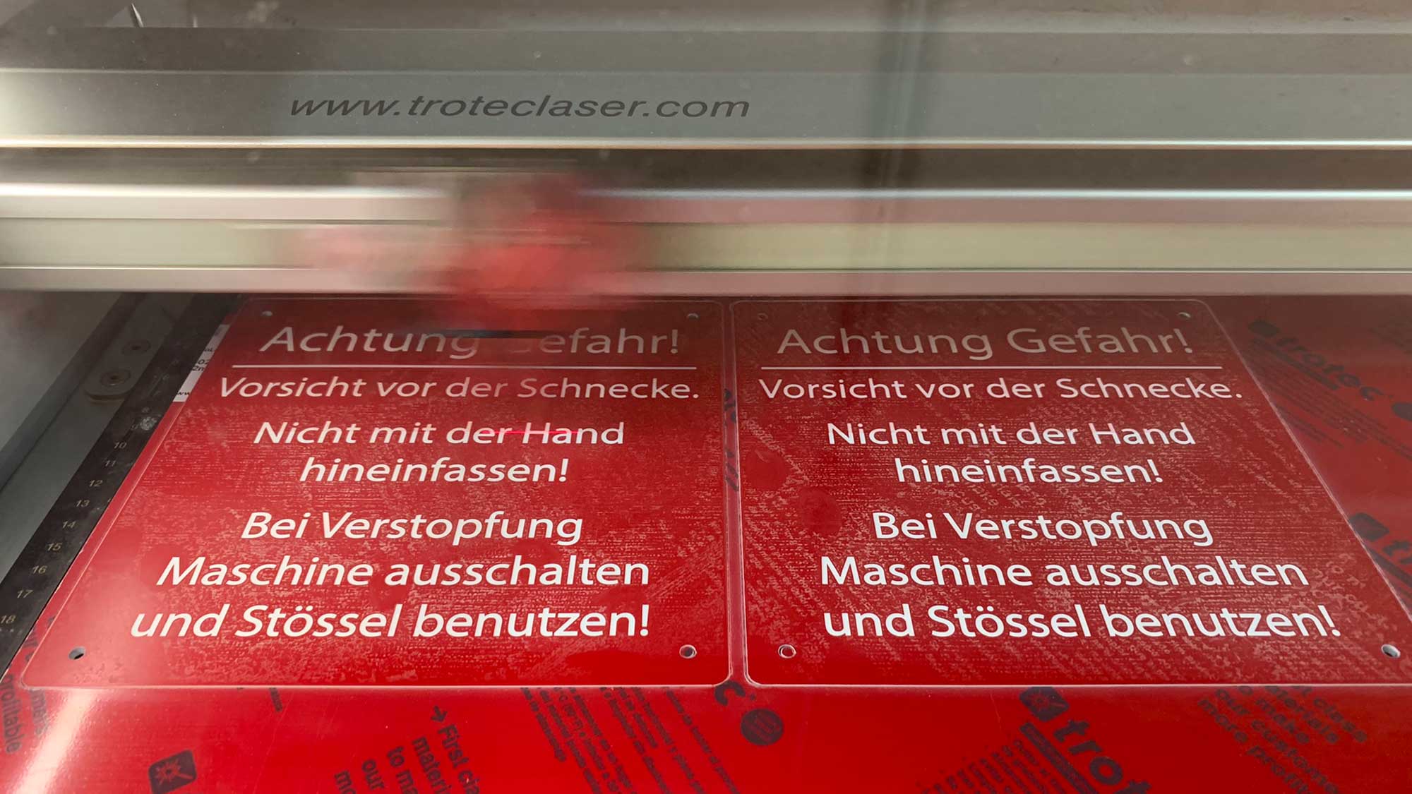 Gravur von zwei roten Kunststoff-Warnschildern mit weißer Schrift in einem CO₂-Laser – Beispiel für industrielle Beschriftung mit hoher Verarbeitungsgeschwindigkeit